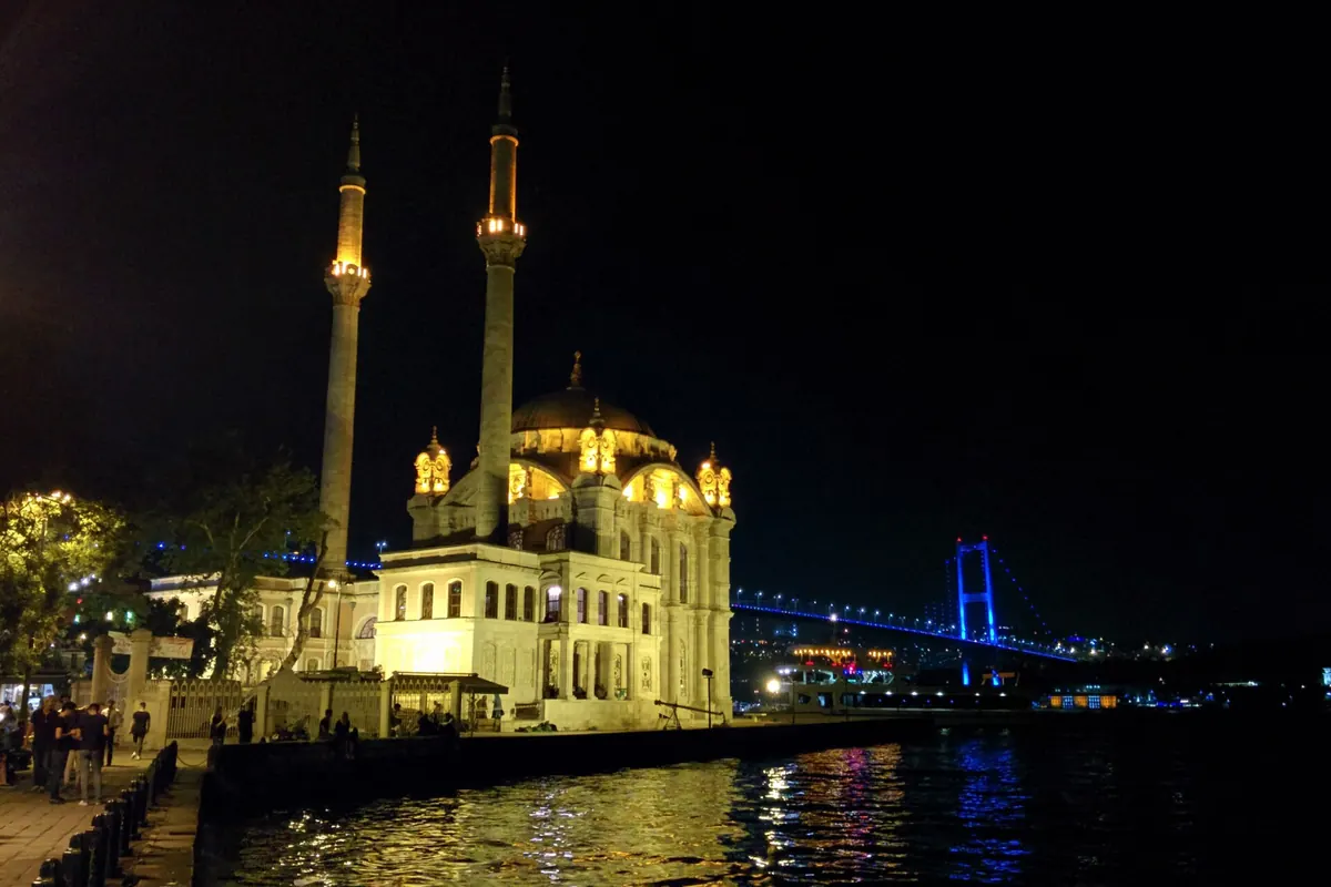 水辺に黄金色にライトアップされたOrtaköy Mosque。細身のミナレットが二本そびえ、背後には青く照らされたボスポラス大橋が浮かびます。